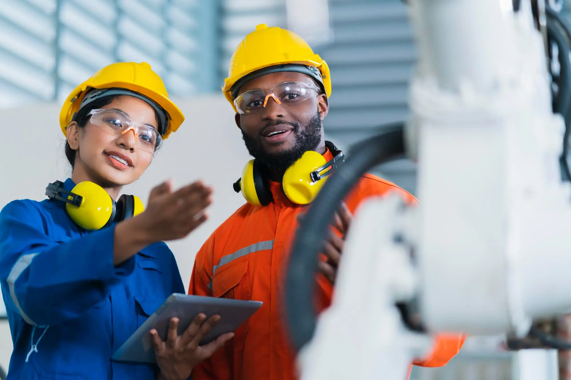 Dois técnicos, uma mulher e um homem, em ambiente industrial, utilizam um tablet enquanto fazem o monitoramento e controle de um sistema. A cena destaca a aplicação da manutenção planejada, com o uso de tecnologia avançada para otimizar processos de prevenção de falhas e reparos.
