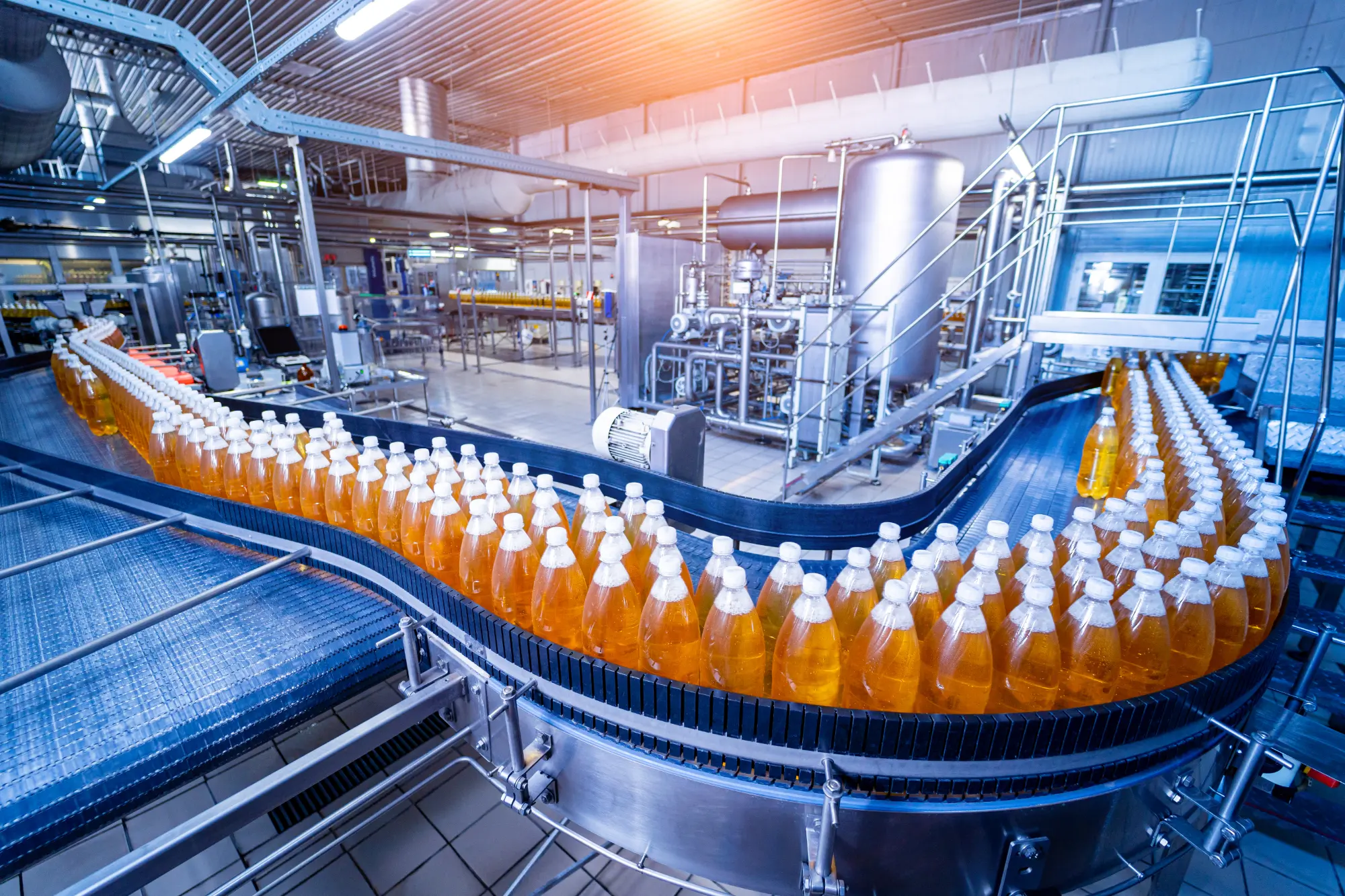 Linha de produção em uma indústria de alimentos e bebidas, com garrafas de uma bebida laranja em uma esteira transportadora. O sistema utiliza válvulas sanitárias para garantir o controle preciso do fluxo e a segurança dos produtos processados.